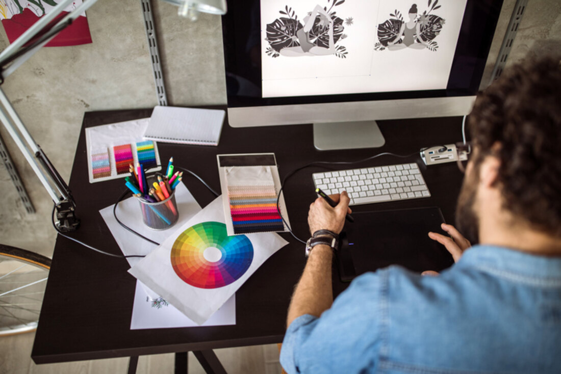 A designer working at a large monitor with colour swatches, printed proofs and pens across the desk