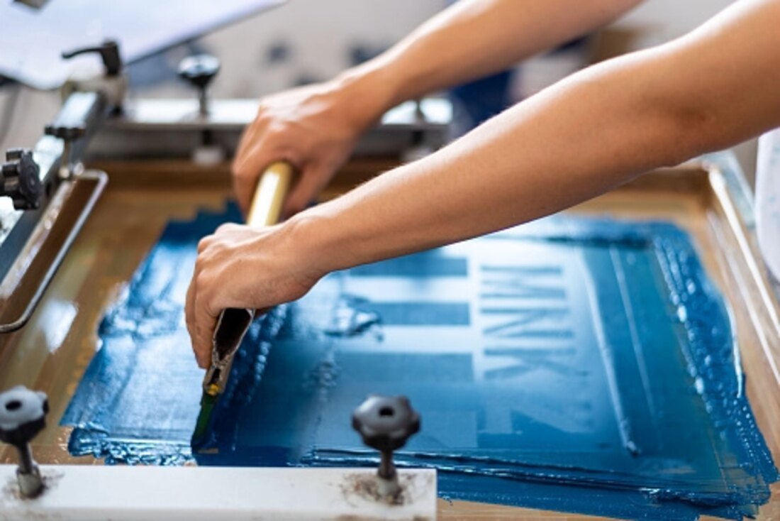 Hands pulling a squeegee across a blue screen on a screen printing press